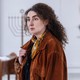 A woman with thick eyebrows and curly hair stands in front of an alter with a visible menorah.
