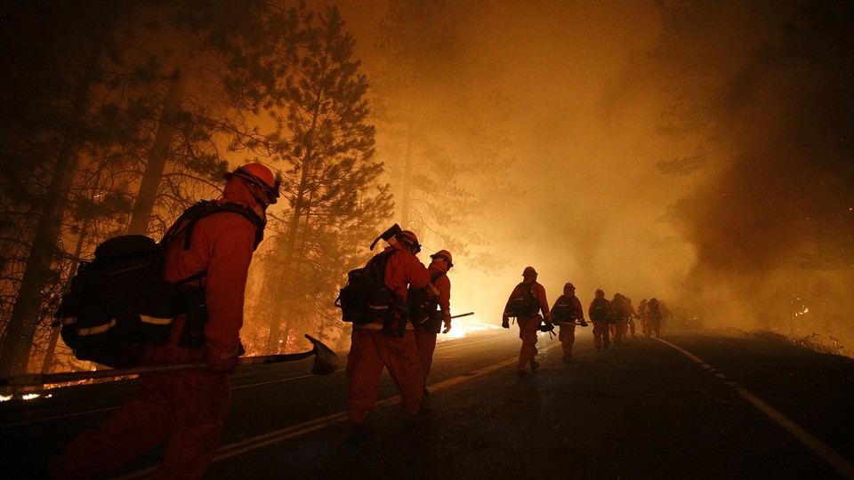 A line of firefighters walks down a road as flames loom in the background.