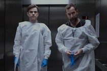 A photo of Patrick Ball and Noah Wyle standing in the elevator with medical gloves on