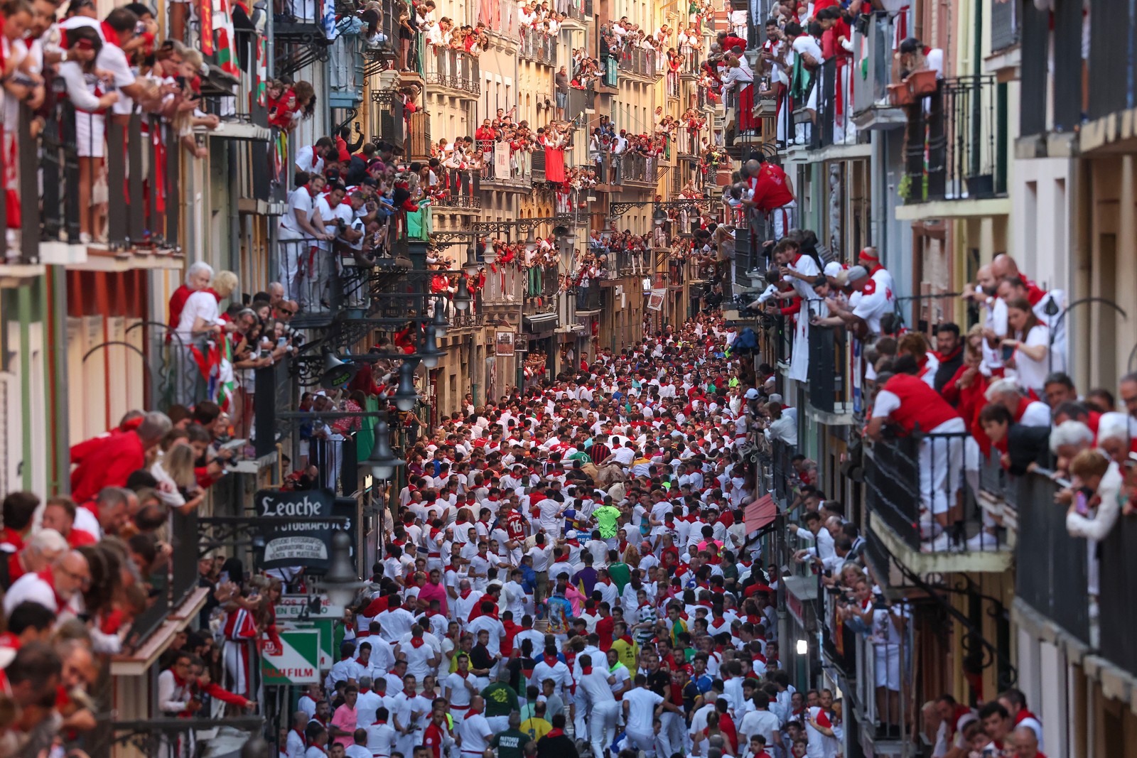 Hundreds of people run in a narrow street as many others stand above on balconies, watching during the 