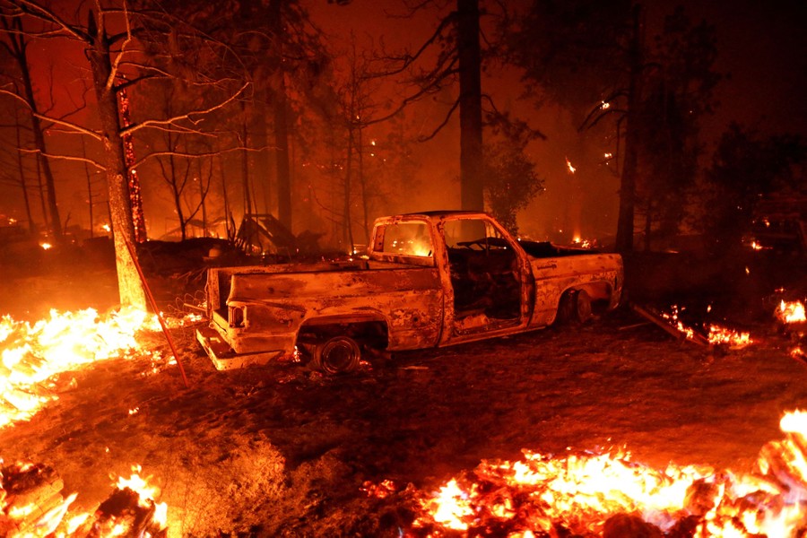 A burned-out truck sits in a forest amid burning trees and scrub.