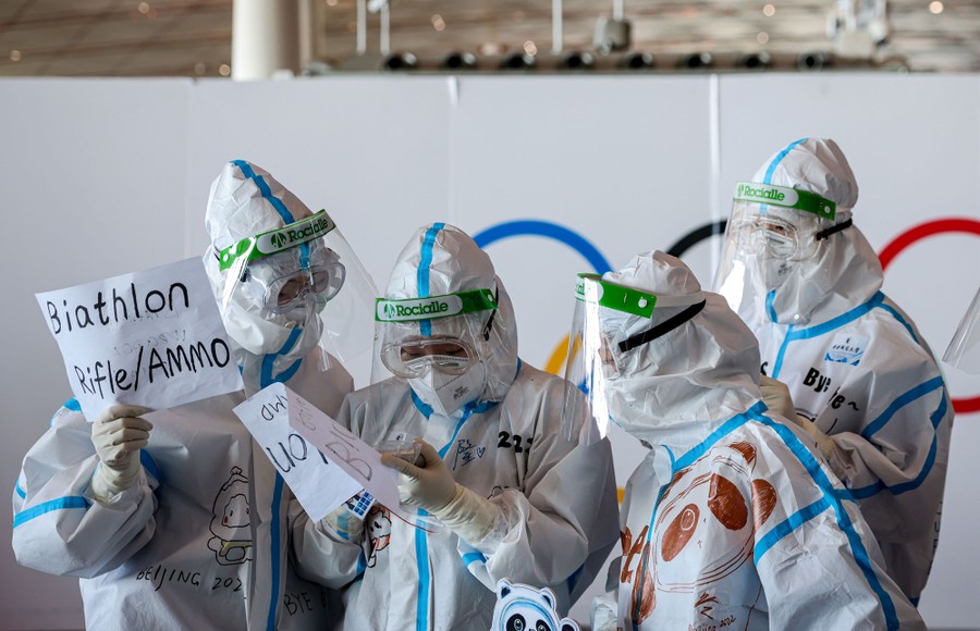Four people in hazmat suits confer while holding handwritten signs to assist Olympic participants.