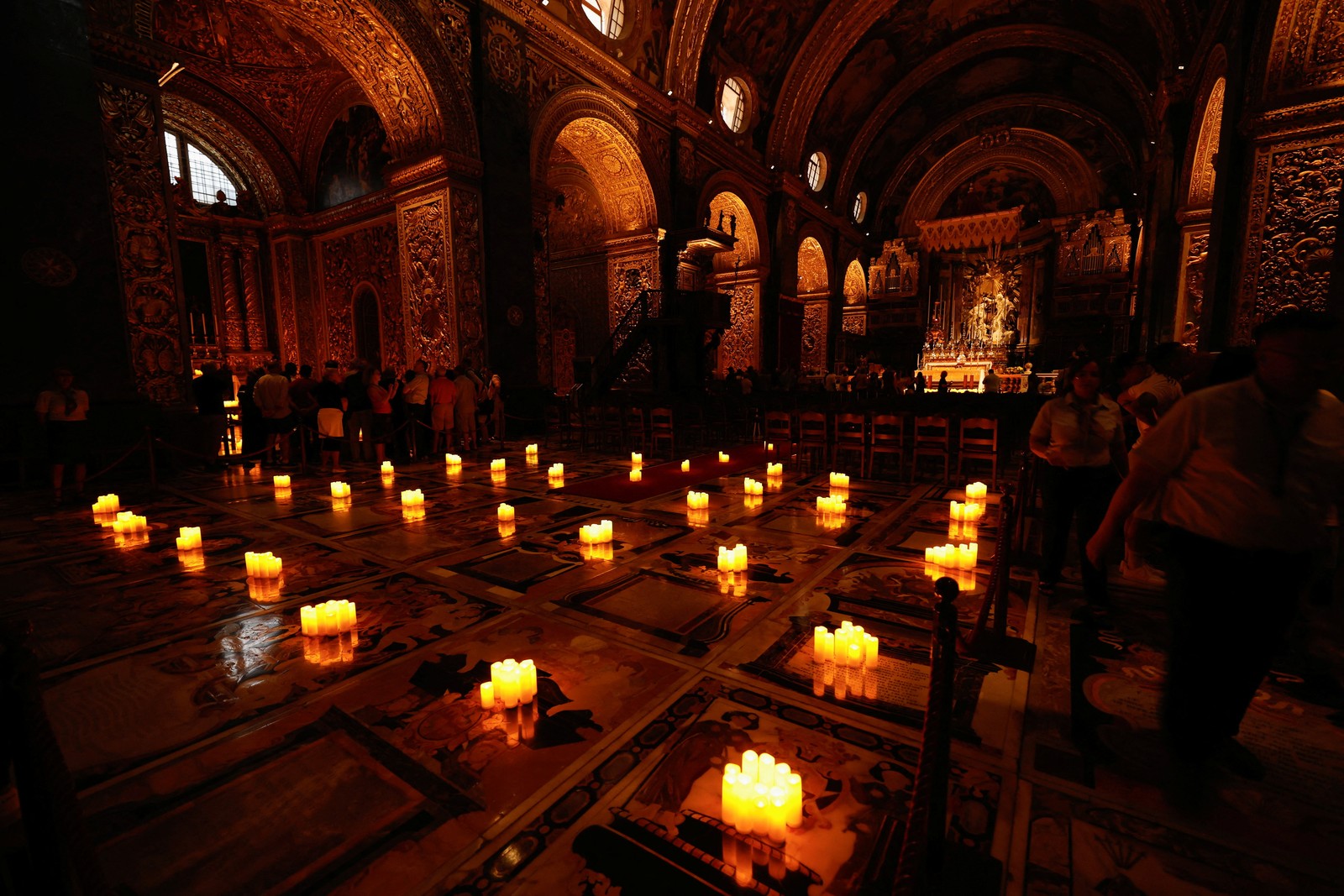 People walk inside a dimly-lit cathedral with many candles set up all around on the floor.