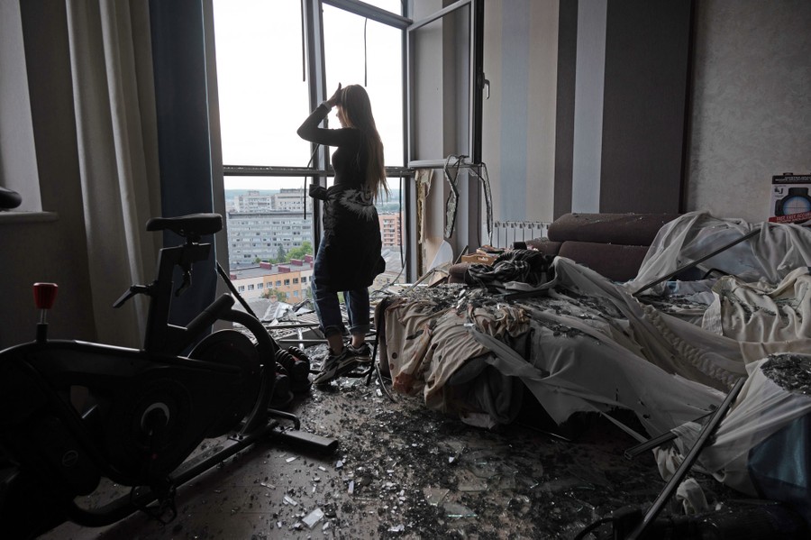 A person stands inside a debris-covered room in an apartment building, looking out broken windows.