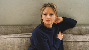 A portrait photograph of the actor Jodie Foster curled up barefoot on a couch, wearing jeans and a navy-blue sweater.