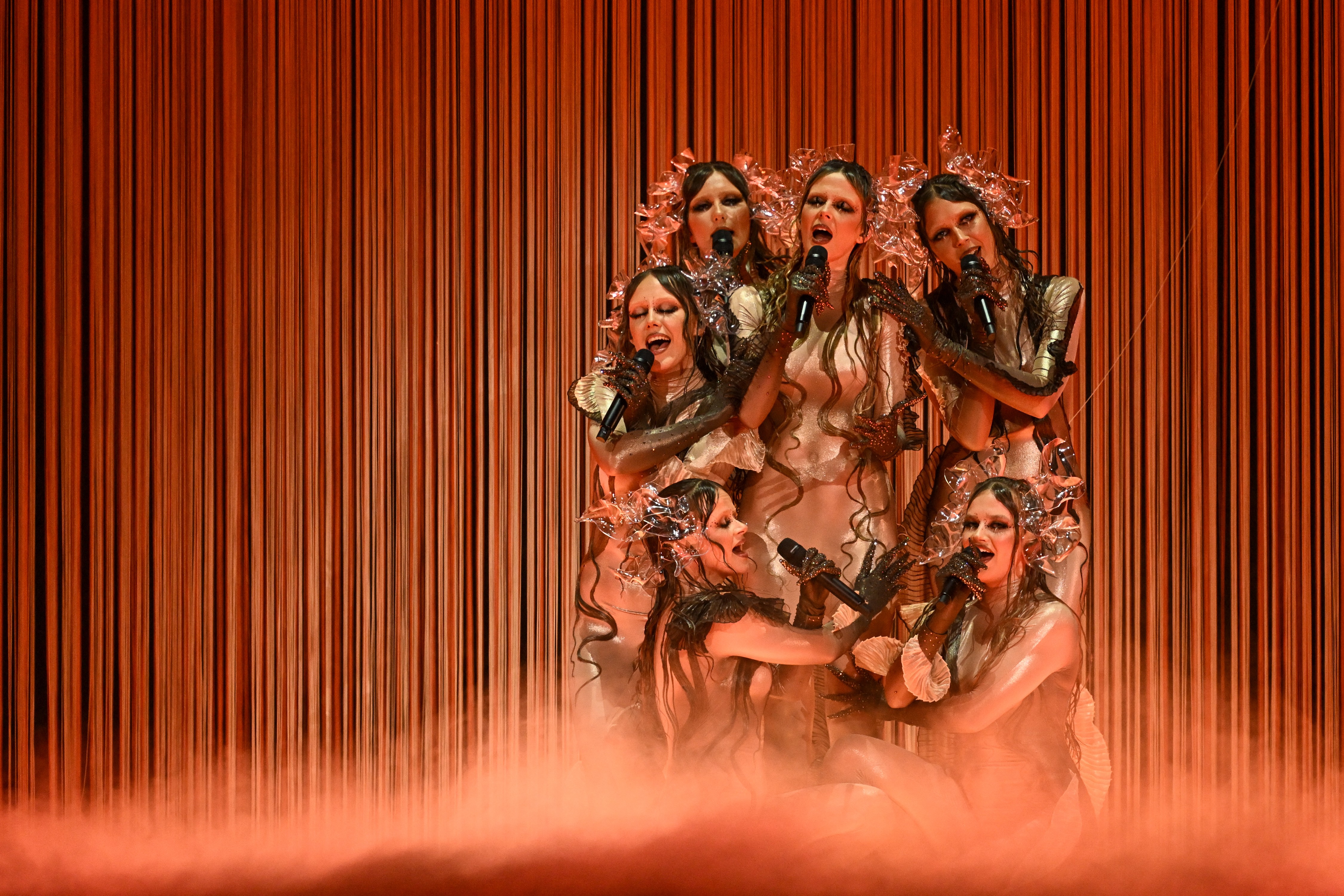 Six costumed performers sing on a stage, clustered together.