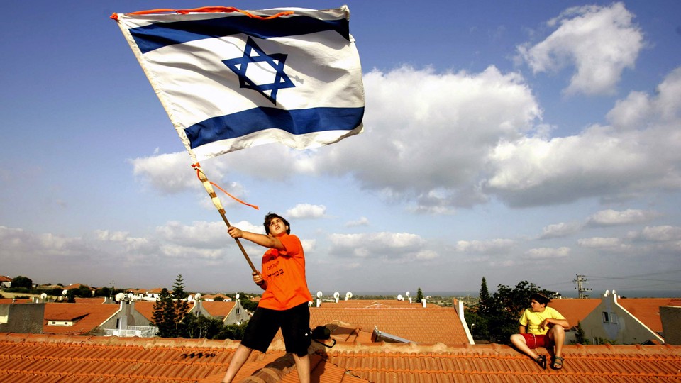 A person waving a large Israeli flag while standing on a roof with a big sky in the background
