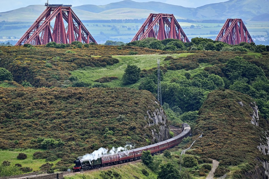A steam train travels down a track with a large bridge visible in the distance.