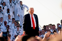 President Donald Trump is shown speaking, with his mouth open, surrounded by uniformed members of the U.S. Navy.