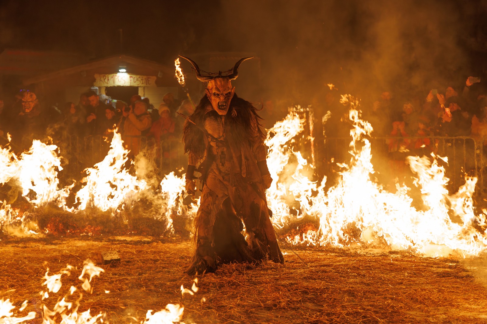 A crowd gathers to watch a person, dressed as Krampus, perform among many small fires.