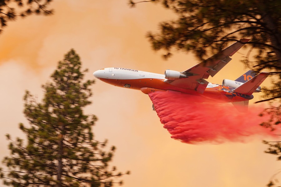 A large aircraft flies close overhead, dropping a red-colored plume of fire retardant onto a forest fire.