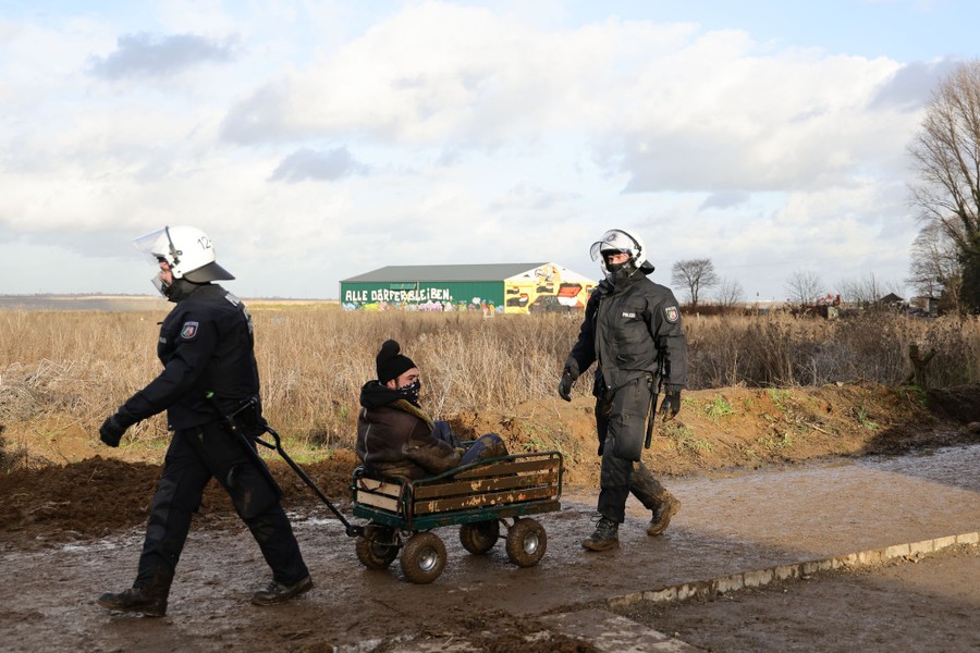 Two police officers walk on a muddy road. One of them pulls a small wagon with a protester seated in it.