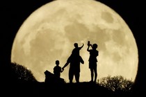 A small group of adults and children standing on a hill is silhouetted by the full moon, seen through a long lens.