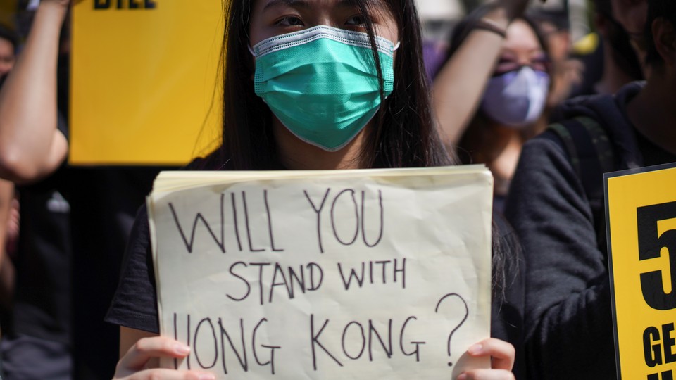 A woman wearing a face mask holds a placard reading, "Will you stand with Hong Kong?"