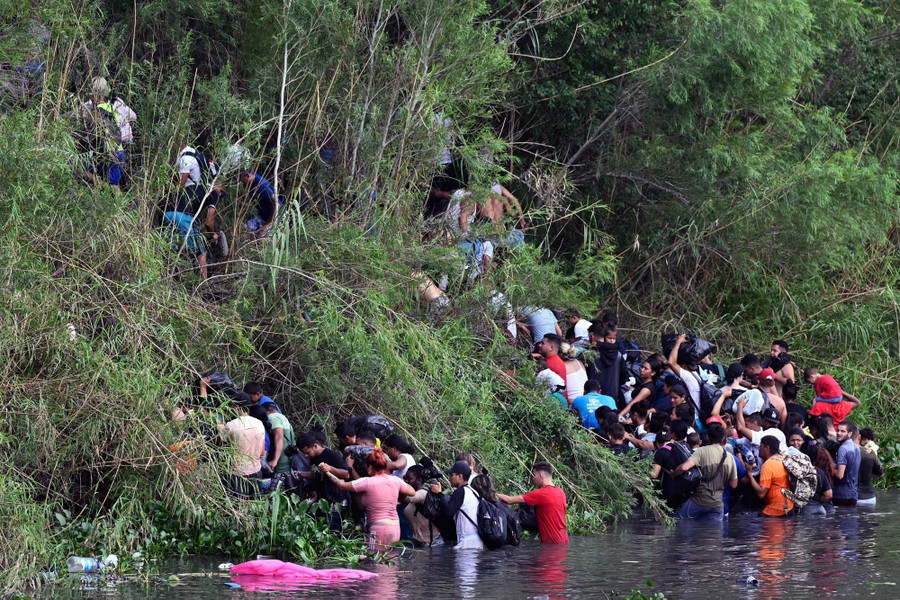 Dozens of people climb a steep hill after wading across a river.