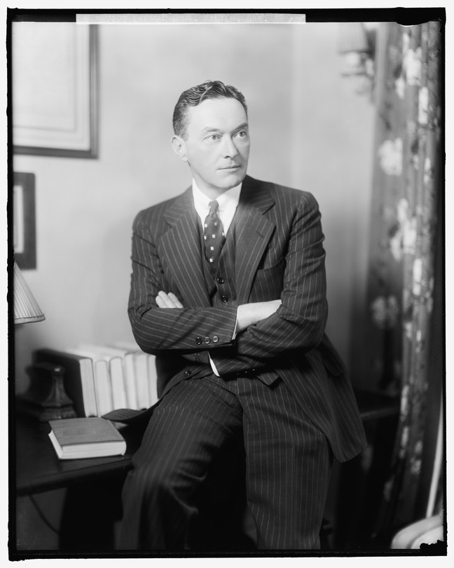 A black and white portrait of Walter Lippmann in a striped suit sitting on a writing desk in front of some books
