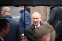 Vladimir Putin stands under an umbrella in the rain, surrounded by people facing the opposite direction.