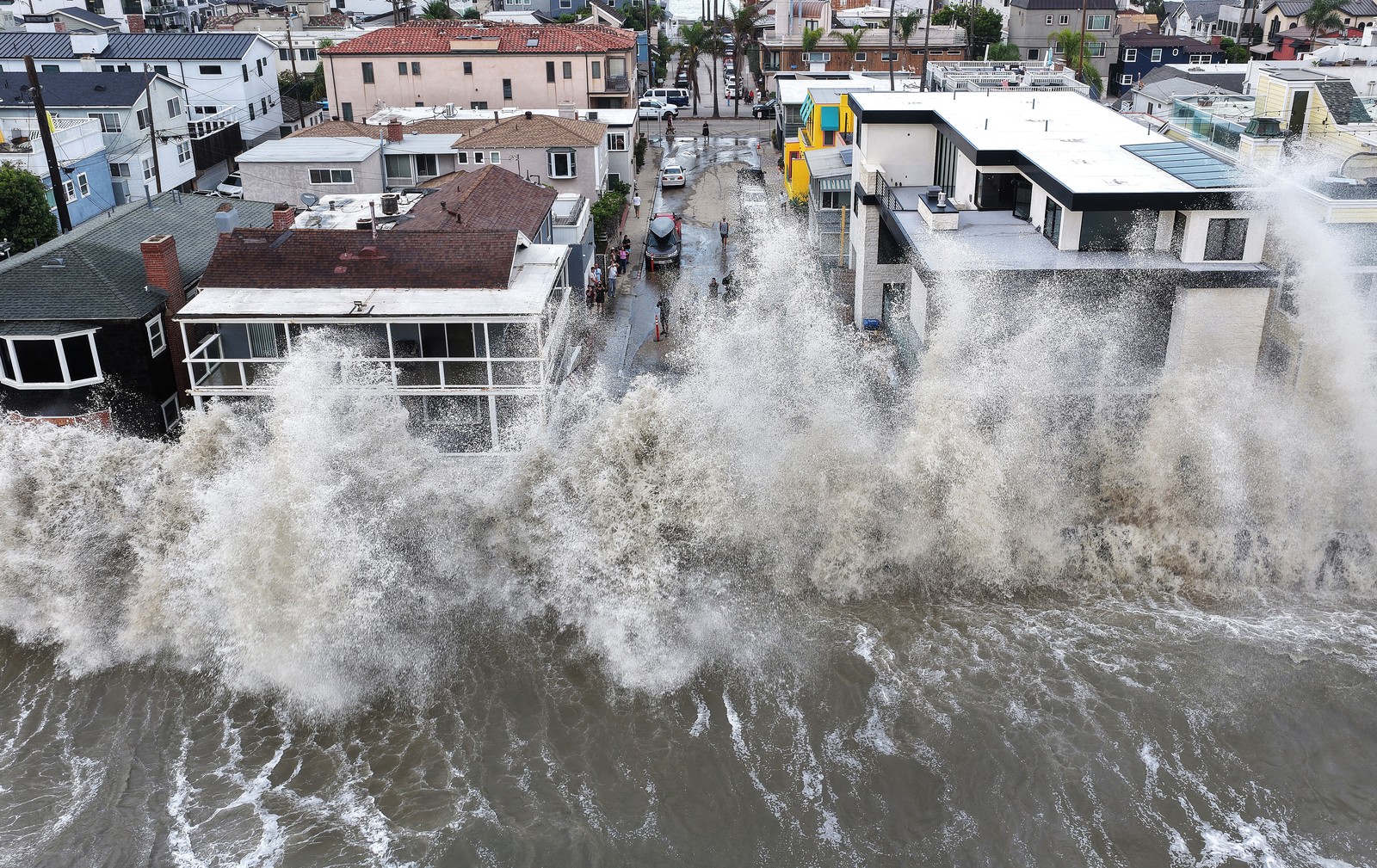An aerial view of a wave crashing against a seawall in front of a residential neighborhood.