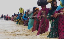 A line of women and men carry bundles through fast-flowing knee-deep floodwater while holding a rope.