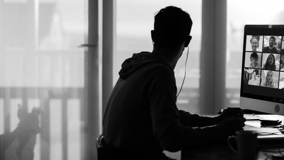 A black-and-white photo of a man working at a desk in his apartment. A Zoom meeting is open on his computer screen, and he seems to be looking out a window.