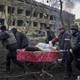 Five men carry a pregnant woman on a stretcher. They are surrounded by destroyed buildings and debris.