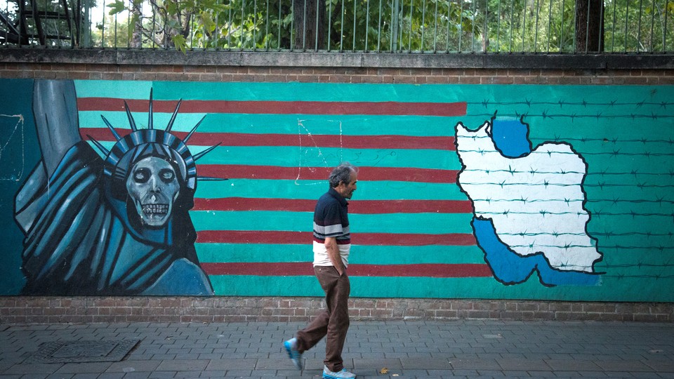 A man in front of an anti-U.S. mural featuring the Statue of Liberty, the American flag, and a map of Iran in barbed wire