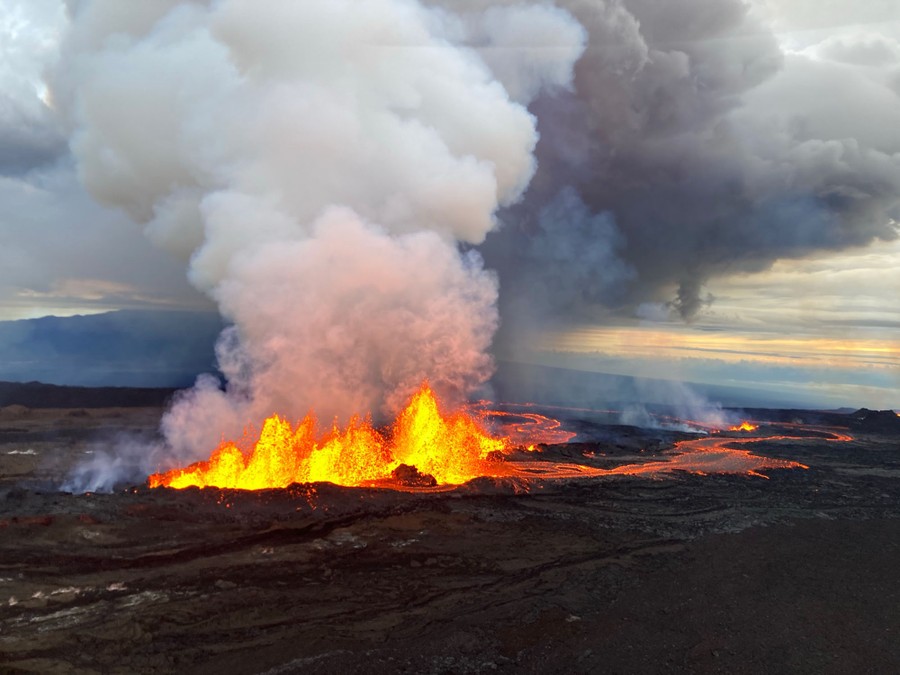 An aerial view of lava spewing into the air from a volcanic fissure, sending up plumes of gas and ash