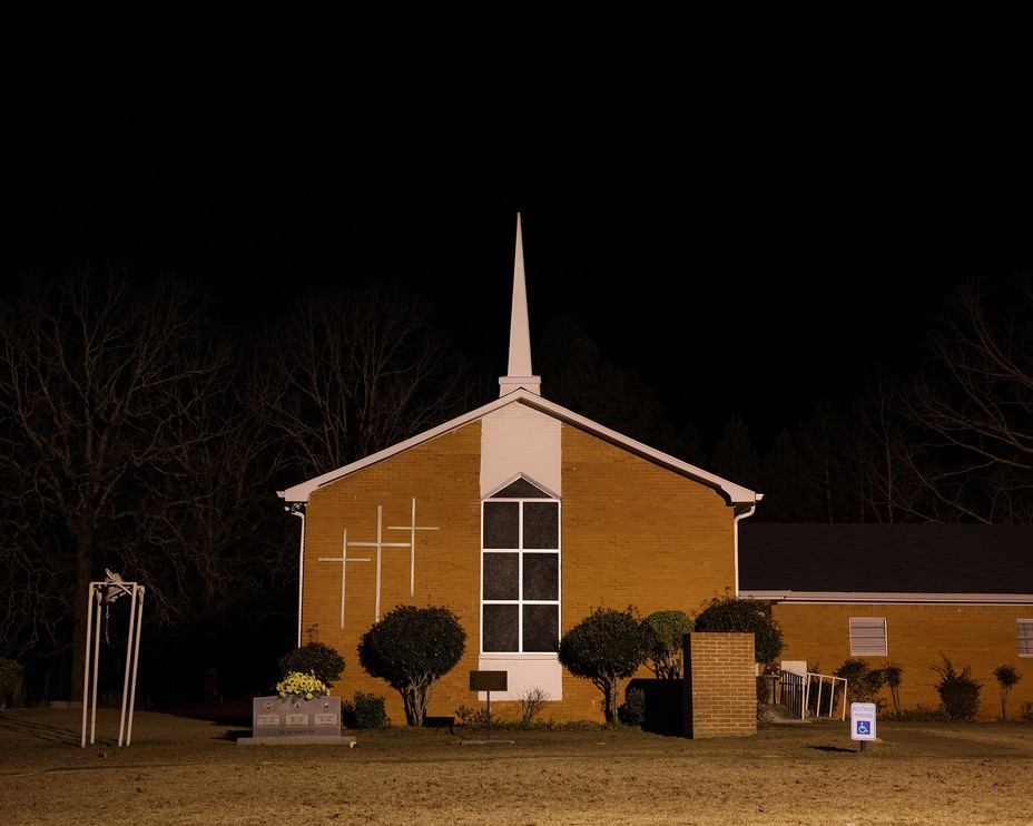 photo at night of a brick church with white steeple and 3 white crosses on the building, with small belltower and memorial marker