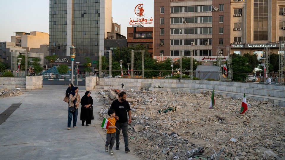 A Iranian family walks by a field of rubble in an urban neighborhood, the young boy carrying an Iranian flag.