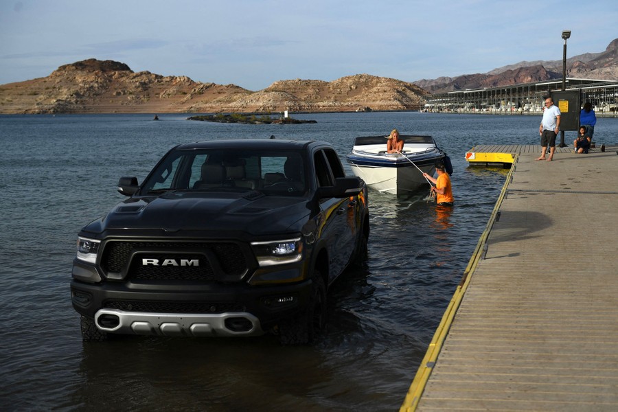 A pickup truck sits in shallow water, alongside a dock waiting to hook up to a small boat.