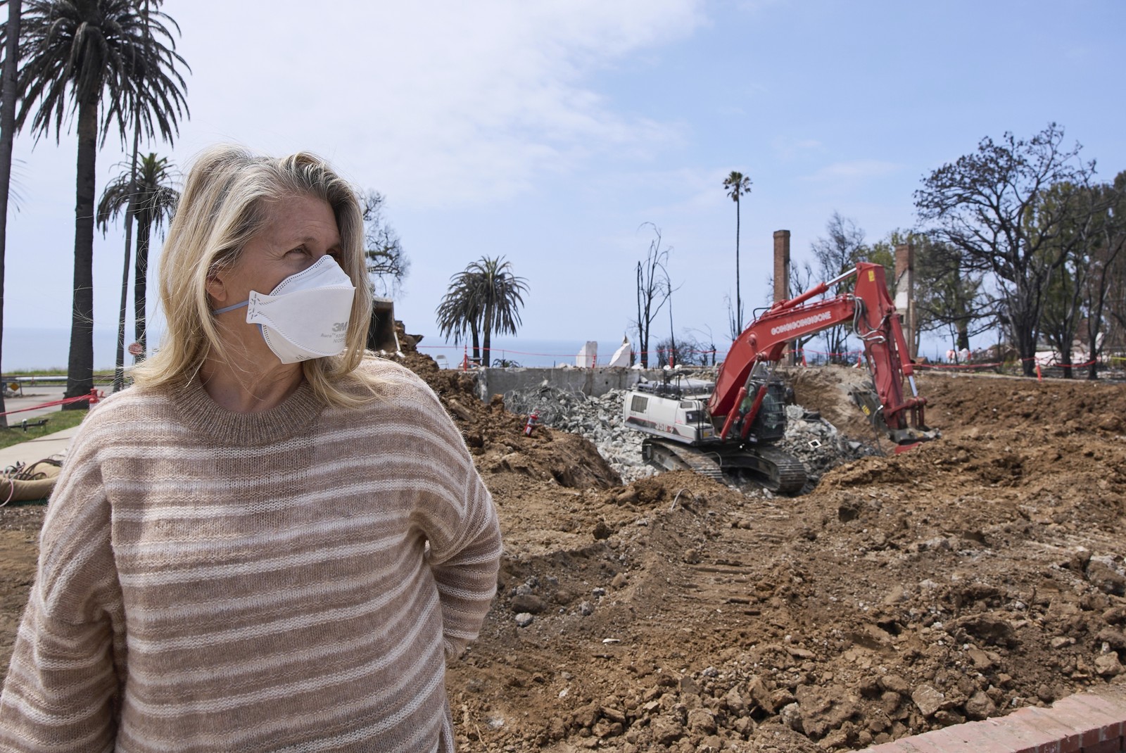 A person wearing a protective face mask stands in front of an excavator, working on a residential lot.
