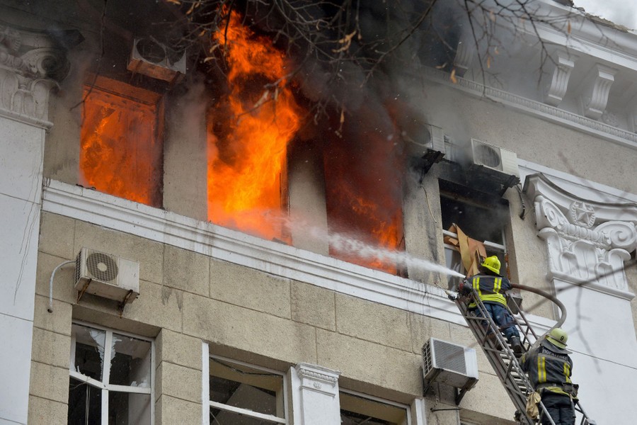 Two firefighters stand atop a long ladder, spraying water into an open window of a tall building where flames are emerging.
