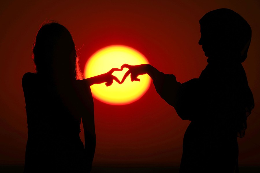 Two young people hold their hands close together, their fingers forming a heart shape, in silhouette against the setting sun.