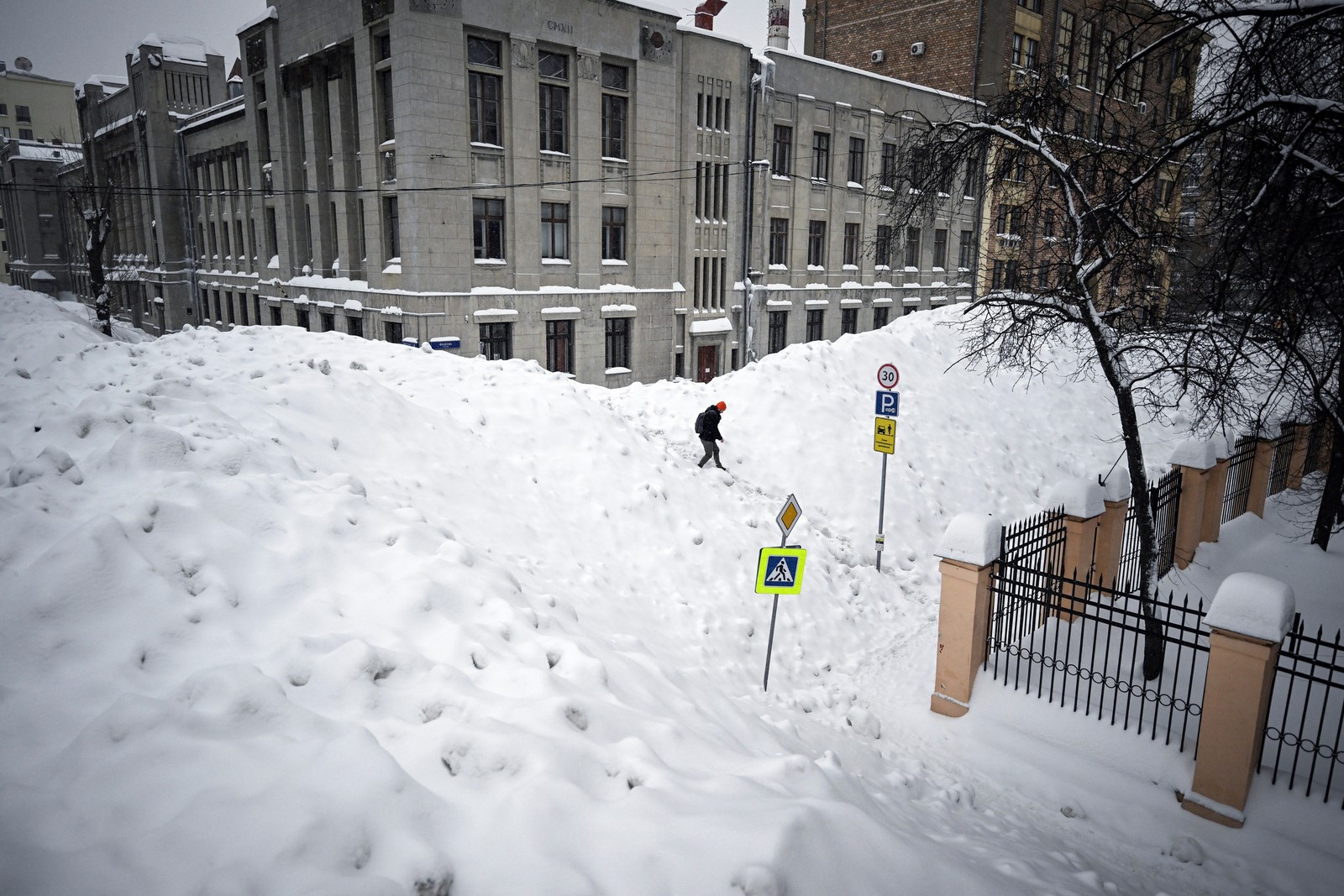 A pedestrian crosses huge mounds of snow on a city street.