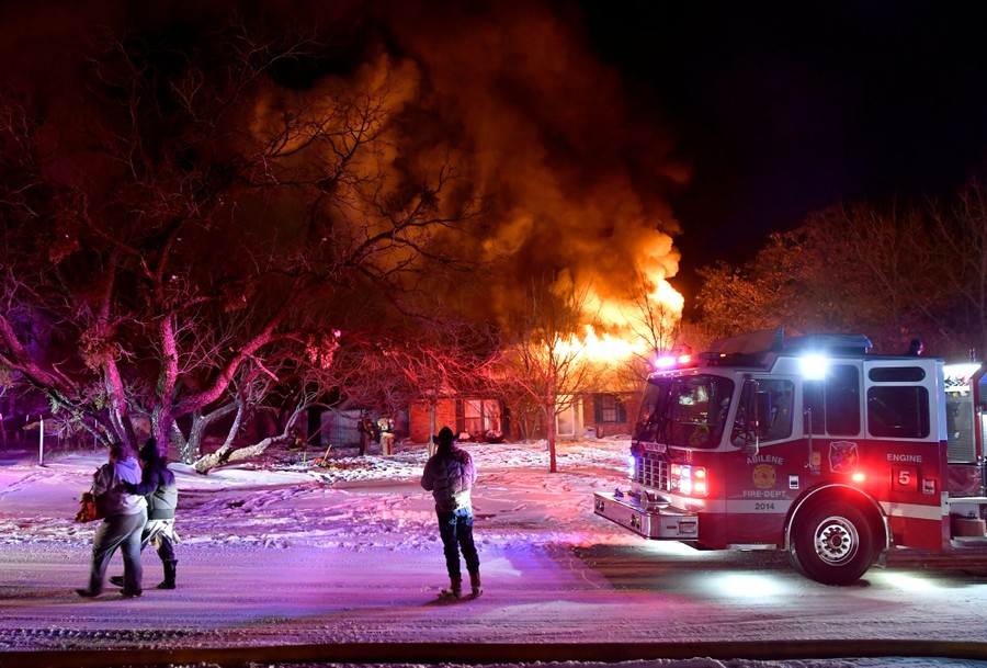 Firefighters stand in a street near a firetruck, as a house burns nearby.