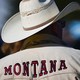A photograph of a man from behind, wearing a white cowboy hat and a white jacket that says MONTANA in red font across the shoulders
