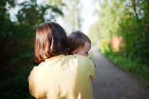 Color photograph of a woman holding a baby, viewed from behind on a wooded path