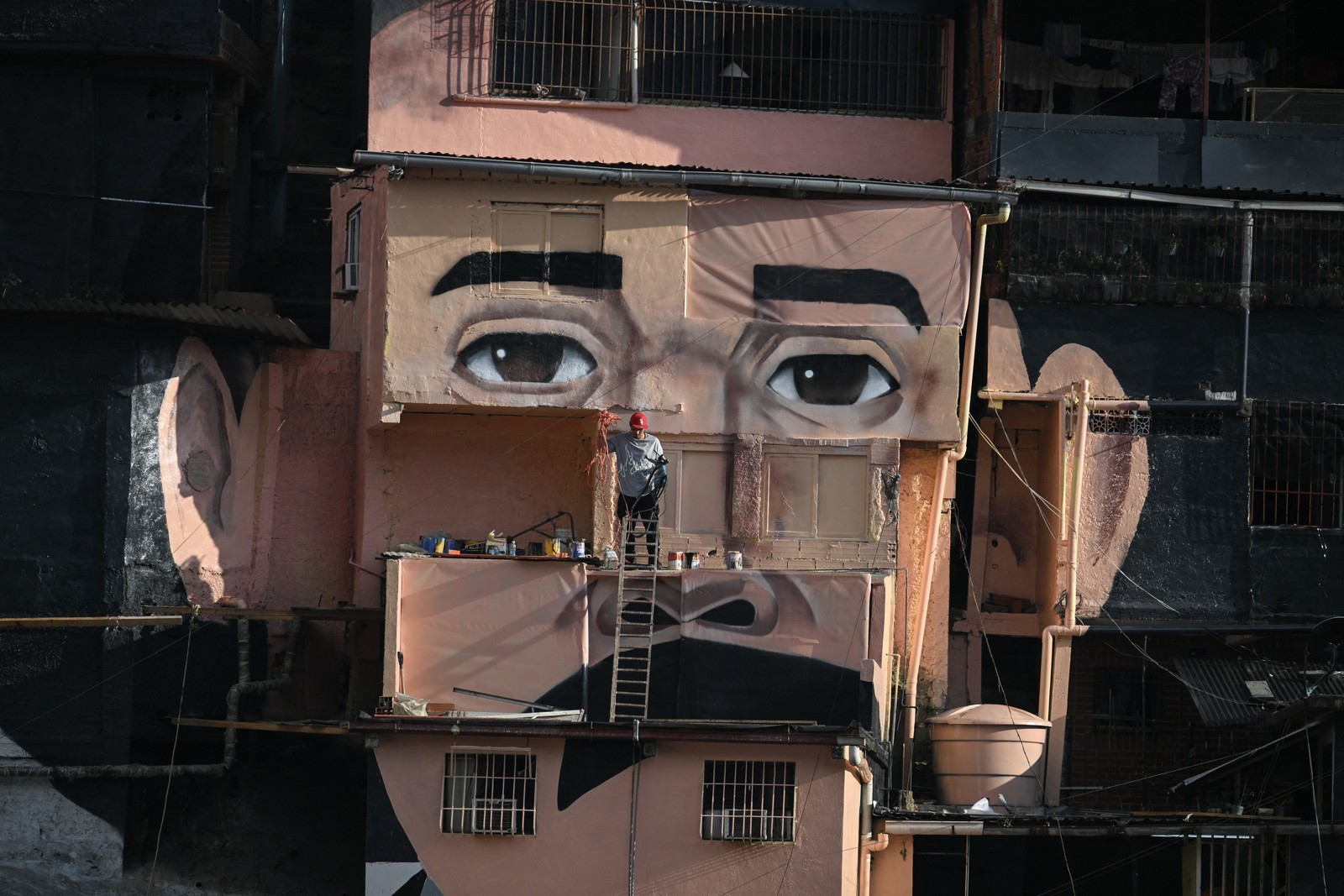 A man works in front of a giant mural of a person's face, painted across several tall building walls.