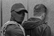 a black and white photo of a masked ICE agent detaining a man in immigration court.