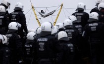 A person sits in a hammock suspended high above the ground from ropes and logs as a large group of riot police gather below.
