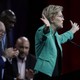 Elizabeth Warren, dressed in an open green jacket, stretches her arms out to her sides as she speaks to an audience. Three men are visible onstage behind her.