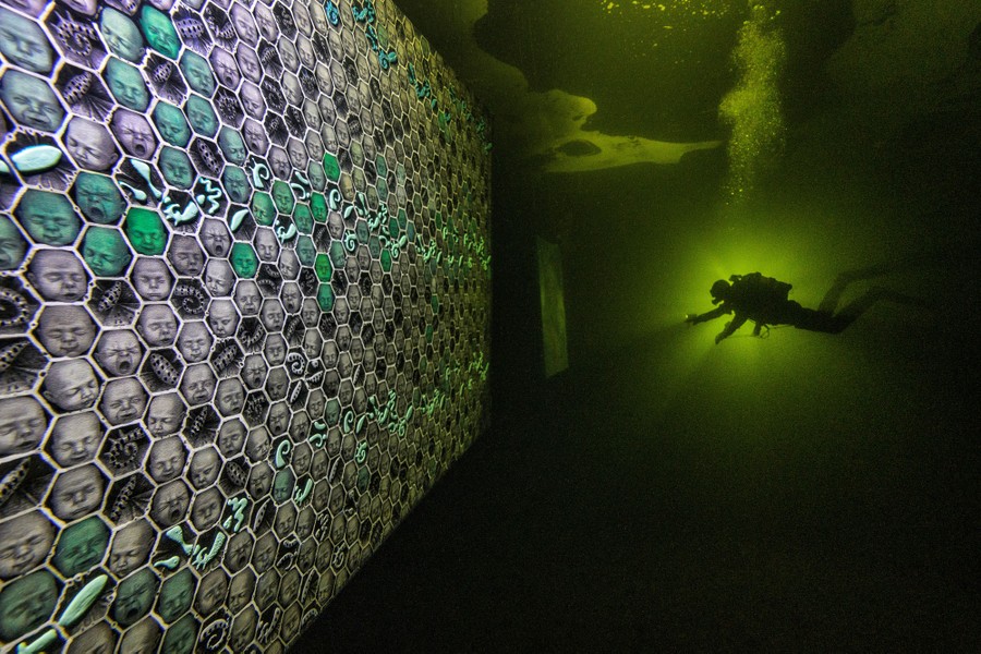 A scuba diver swims in dark water, shining a light toward large underwater panels covered with artwork - images of baby faces.