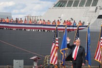 Trump waves in front of flags and a large military ship filled with men taking photos of him