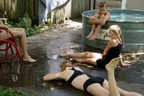 four kids sitting around outside in bathing suits, one sitting at the edge of a pool, two on the ground, one in a red chair