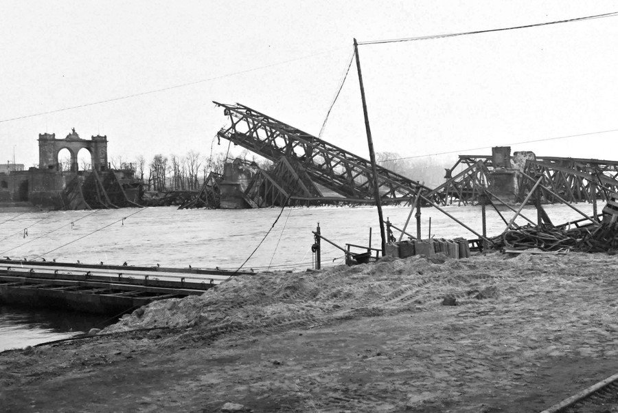 The wreckage of a huge steel-truss road and rail bridge lies across a broad river.