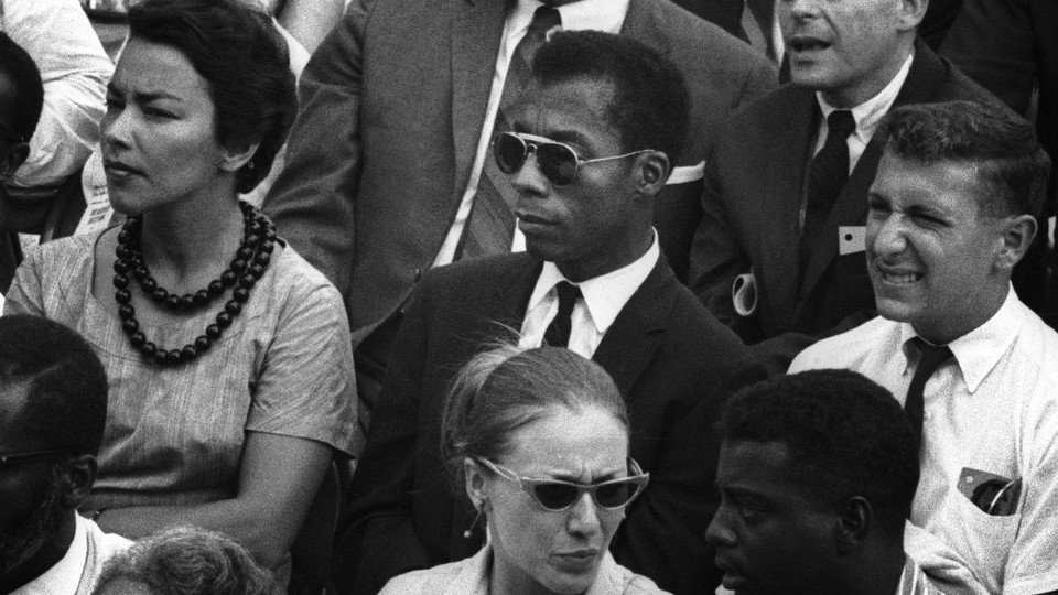 James Baldwin, seated among other people, wearing sunglasses and a suit and tie.