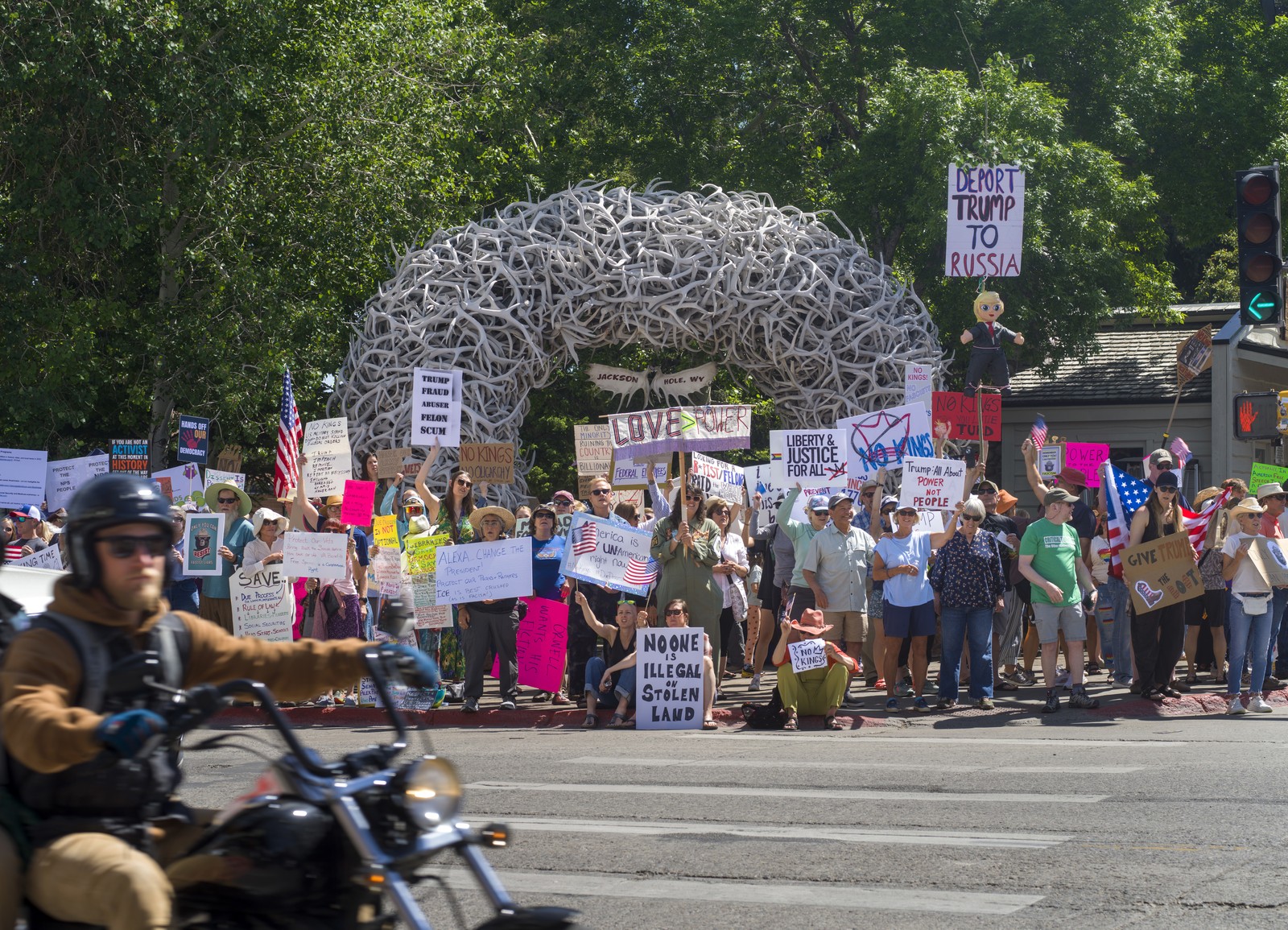 Demonstrators hold signs while standing on a street corner in front of an arch built out of antlers.
