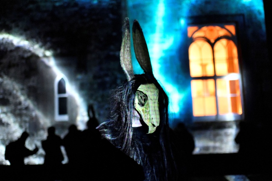A scary costumed character walks in front of a castle wall that has images projected on it.