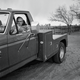 black-and-white photo of author sitting in driver's seat of large dually truck on dirt road with fence and tree in background