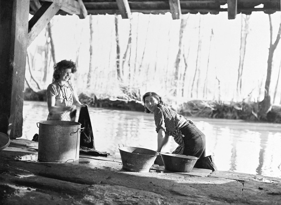 Two women kneel beside a stream, under an awning, washing clothes by hand.